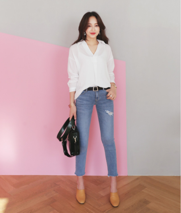 Người phụ nữ mặc áo sơ mi trắng và quần jeans, đang cầm túi xách và đứng trước bức tường màu hồng.